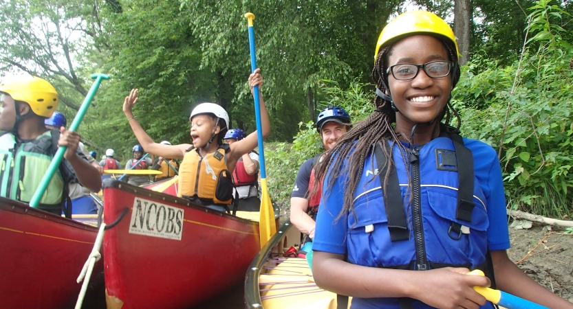 A group of students wearing safety gear sit in canoes and smile. They appear to be near the shore, close to trees.